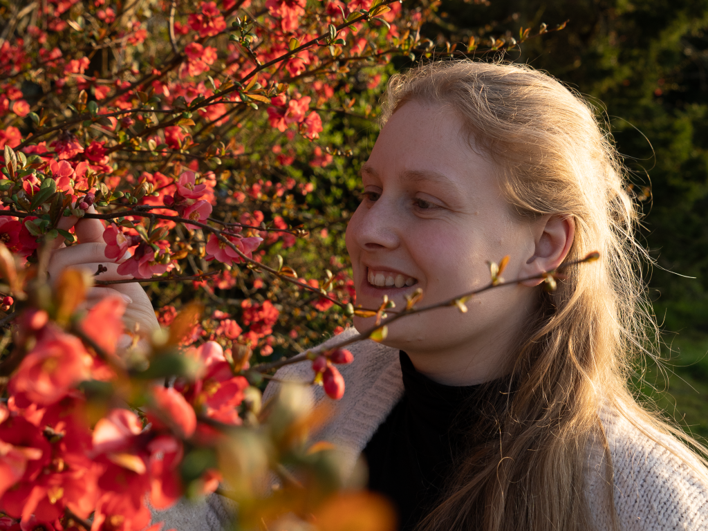 Eine blonde Frau lächelt und hebt mit einer Hand einen Zweig von dem vor ihr liegenden Busch. Der Busch hat rote Blüten und nimmt die ganze linke Bildhäfte ein. Von hinten scheint die Sonne auf das blonde Haar der Frau und lässt es fast weiß wirken. Sie trägt einen schwarzen Rollkragenpullover und darüber eine Beige Strickjacke.