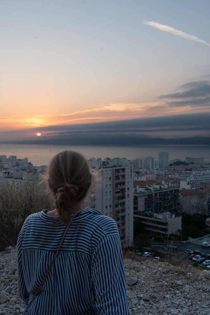 Eine junge Frau mit blonden Haaren in einer gestreiften Bluse ist von hinten zu sehen. Sie trägt einen Dutt. Sie blickt in Richtung eines Sonnenuntergangs. Darunter ist eine Stadt sichtbar.