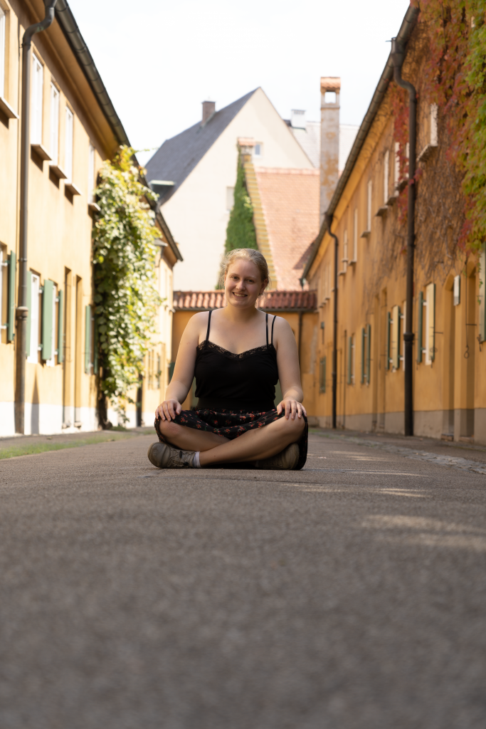 Eine blonde Frau sitzt im schwarzen Rock und Top im Schneidersitzt in der Mitte einer Gasse. Beige Häuser sind auf beiden Seiten des Wegs sichtbar.