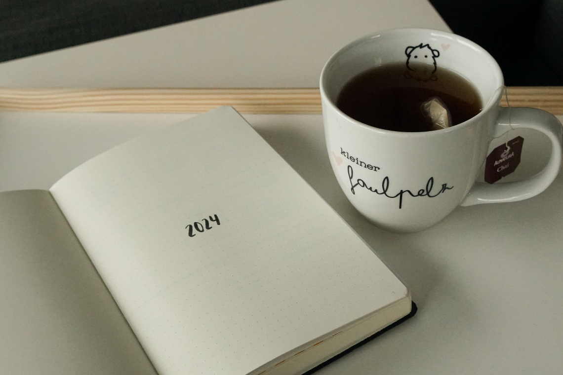 Auf einem Tablet liegt ein aufgeschlagenes Notizbuch mit der Aufschrift "2024". Daneben steht eine Tasse mit Tee und der Aufschrift "kleiner Faulpelz".