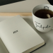 Auf einem Tablet liegt ein aufgeschlagenes Notizbuch mit der Aufschrift "2024". Daneben steht eine Tasse mit Tee und der Aufschrift "kleiner Faulpelz".