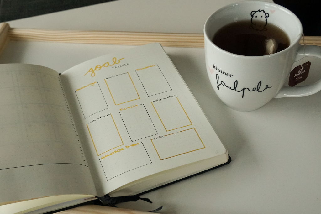 Auf einem Tablet liegt ein aufgeschlagenes Notizbuch mit der Aufschrift "Goal Tracker" und acht Kästchen. Daneben steht eine Tasse mit Tee und der Aufschrift "kleiner Faulpelz".