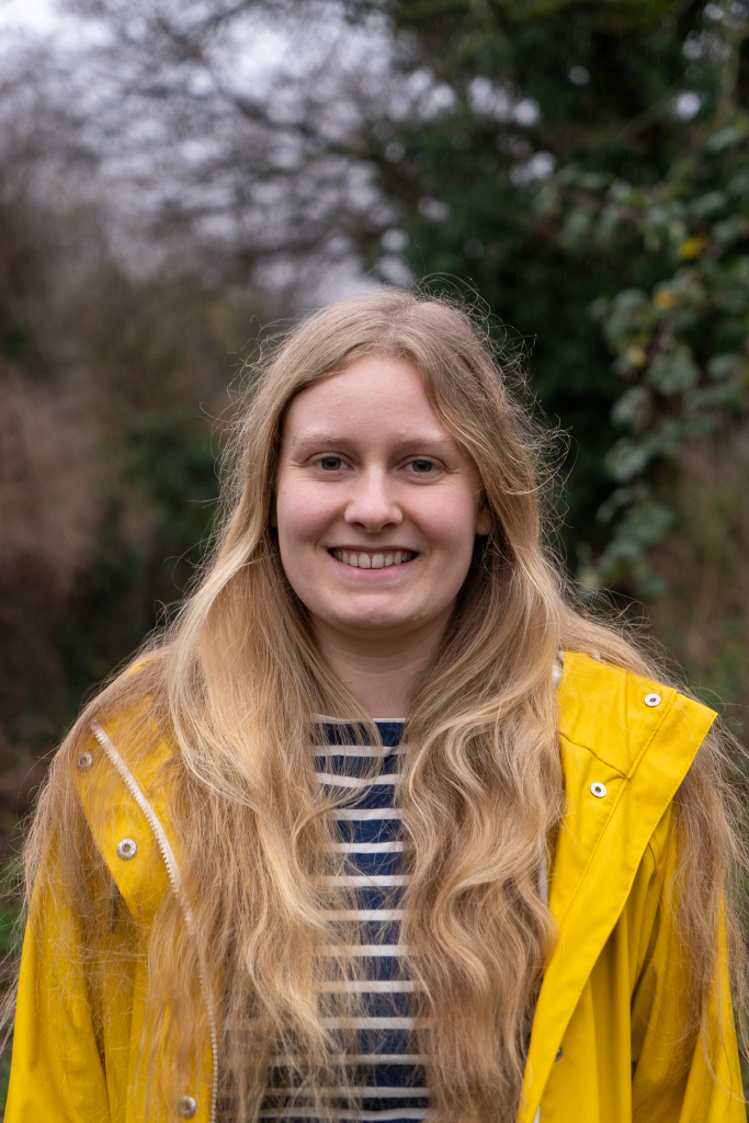 Das Profilbild einer jungen Frau mit langen blonden Haaren. Sie trägt ein blau-weiß gestreiftes Shirt und eine gelbe Regenjacke. Im Hintergrund erkennt man verschommene Pflanzen.