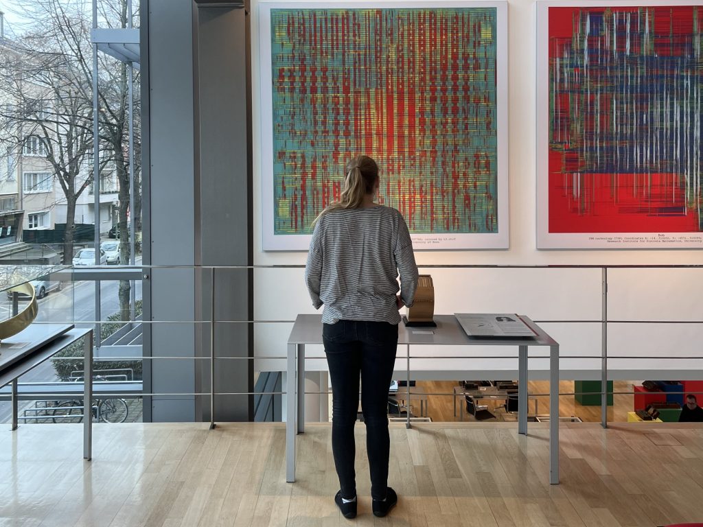 Eine Frau steht an einer Balustrade und betrachtet ein Bild. Sie trägt ein schwarze-weiß gestreiftes Langarmshirt und eine schwarze Hose. Ihre blonden Haare sind in einem Pferdeschwanz. Das betrachtete Bild scheint eine abstrakte Anordnung von gelben und vorgegangen Quadraten unterbrochen von türkisen Flächen zu sein.