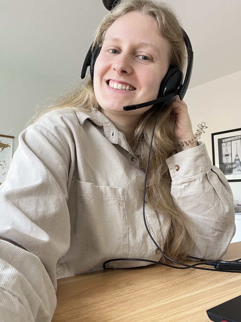 Eine blonde Frau mit schwarzem Headset lächelt die Kamera. Sie trägt ein beiges Kordoberteil.