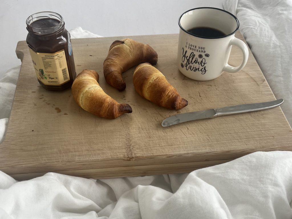 Drei Croissants liegen auf einem Schneidebrett. Ebenfalls darauf befinden sich ein Messer, ein Glas mit Nussnougat-Creme und eine Tasse mit Kaffe. Auf der Tasse steht "I love you a thousend yellow daisies". Das Brett steht auf einem Bett mit weißen Lacken und weißer Bettdecke.