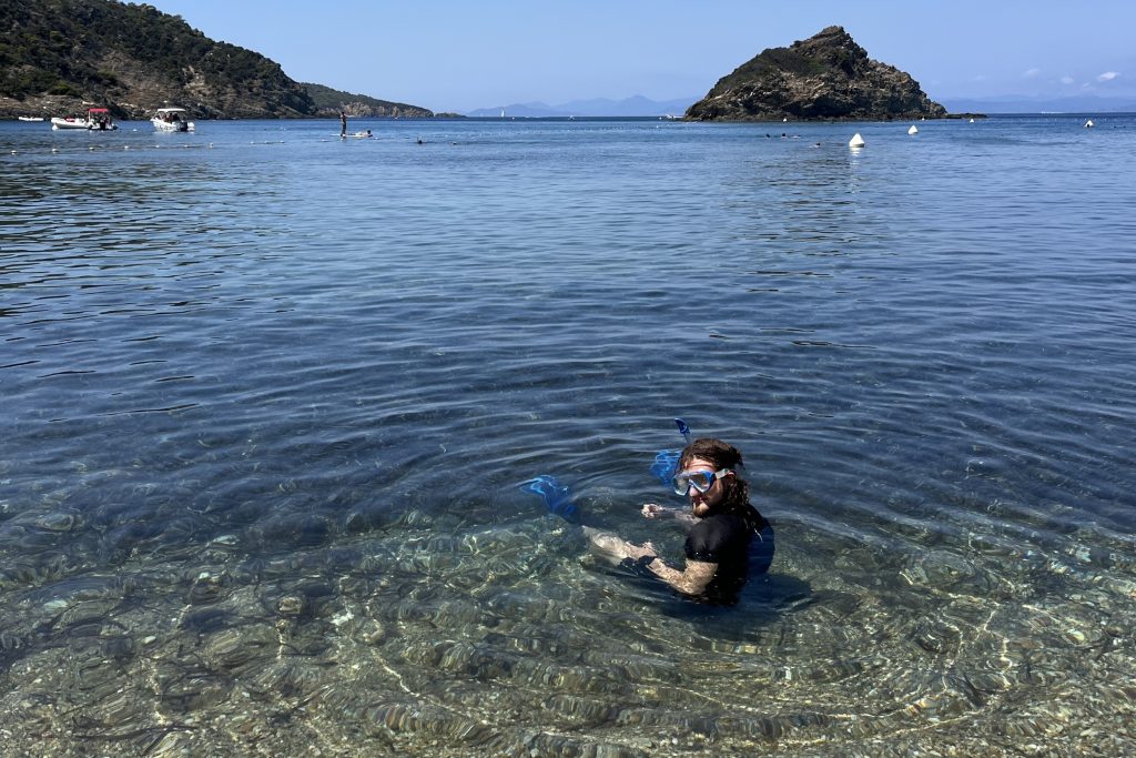 Ein Mann mit dunklen Haaren sitzt im Wasser. Er trägt eine Taucherbrille, einen Schnorchel und blaue Flossen, sowie schwarze Kleidung. Um ihn herum bilden sich Ringe im Wasser. Im Hintegrund ist ein Hügel im Wasser zu sehen sowie ein paar weiße Bojen und kleine Boote.