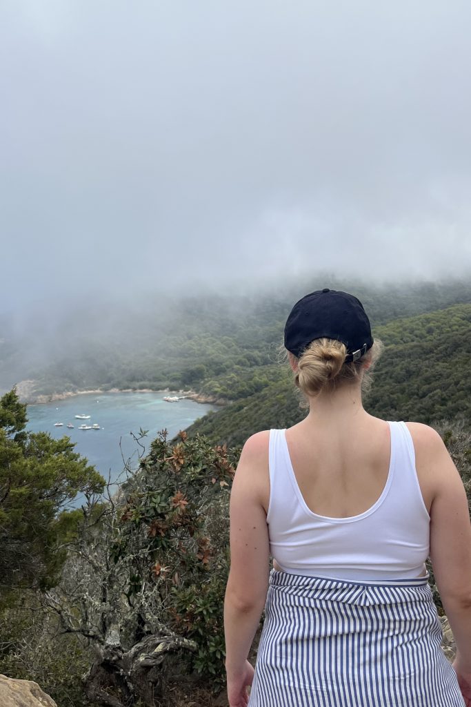 Eine junge Frau mit blondem Dutt und blauer Cap ist von hinten zu sehen. Sie trägt ein weißes Top und ein gestreiftes Oberteil, das sie um die Hüfte gebunden hat. Sie blick in Richtung einer Bucht zwischen grünen Hugeln. Die obere Bildhäfte ist mit Nebel gefüllt.