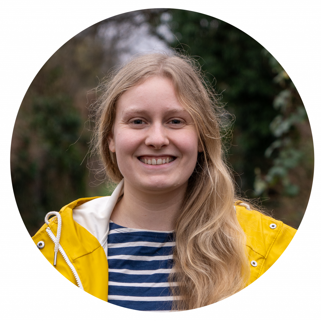 Das Profilbild einer jungen Frau mit langen blonden Haaren, die ihr locker über die linke Schultern fallen. Sie trägt ein blau-weiß gestreiftes Shirt und eine gelbe Regenjacke. Im Hintergrund erkennt man verschommene Pflanzen. 