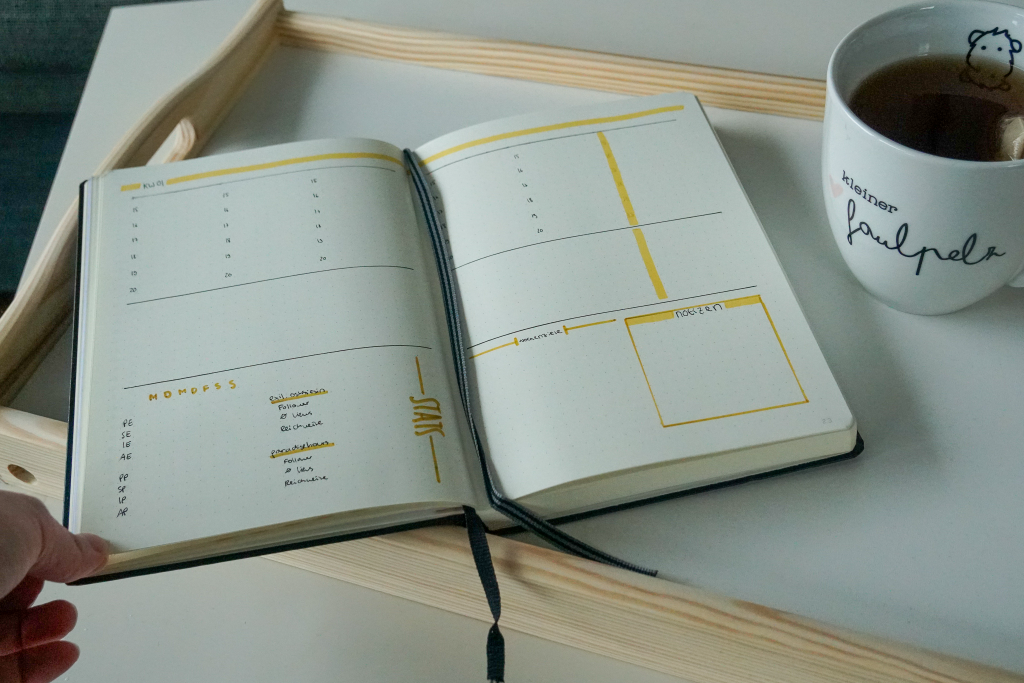 Auf dem Bild ist ein Notizbuch vor weißem Hintergrund zu sehen. Es ist aufgeschlagen. Auf den Seiten erkennt man eine selbst gemalte Kalenderwoche mit einem Kasten für Wochenziele. Neben dem Notizbuch ist ein Tee zu erkennen.