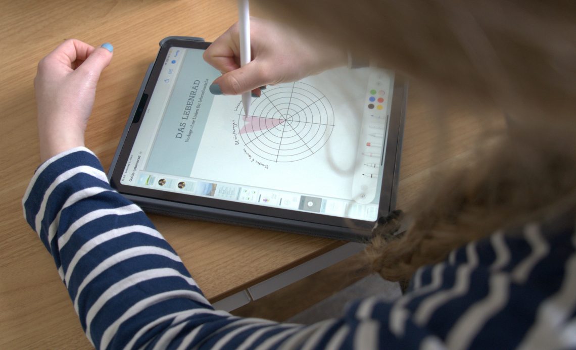 Die betrachtende Person schaut einer blonden Frau über die Schulter. Sie trägt ein blau-weiß gestreiftes Shirt. Vor ihr liegt ein Tablet auf einem Holztisch. Darauf ist ein Tortendiagramm zu sehen, dass sie mit pinker Farbe ausmalt.