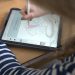 Die betrachtende Person schaut einer blonden Frau über die Schulter. Sie trägt ein blau-weiß gestreiftes Shirt. Vor ihr liegt ein Tablet auf einem Holztisch. Darauf ist ein Tortendiagramm zu sehen, dass sie mit pinker Farbe ausmalt.