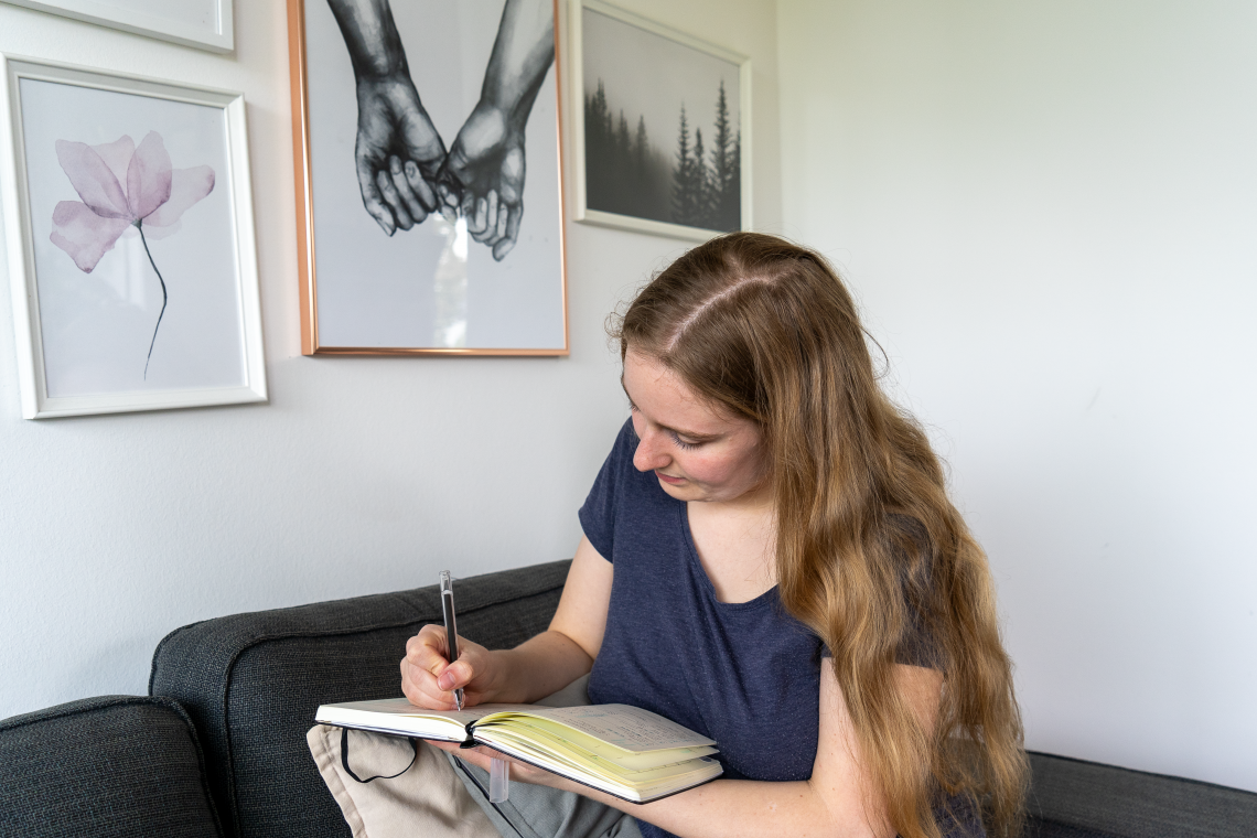 Auf dem Bild sieht man eine blonde Frau mit langen Haaren. Sie trägt ein blaues T-Shirt und schreibt i nein Notizbuch. Im Hintergrund sieht man ein paar Bilder an der Wand hängen.
