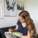 Auf dem Bild sieht man eine blonde Frau mit langen Haaren. Sie trägt ein blaues T-Shirt und schreibt i nein Notizbuch. Im Hintergrund sieht man ein paar Bilder an der Wand hängen.