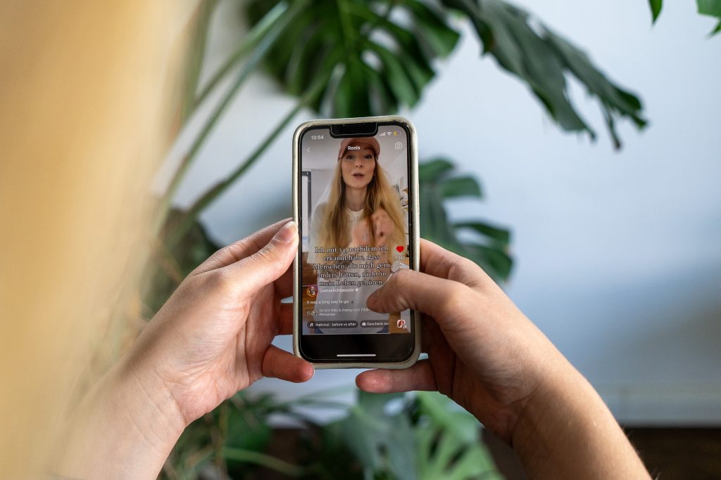 Man schaut einer Frau von hinten über die Schulter. Sie hält ein Handy in der Hand auf dem ein Reel zu sehen ist.
