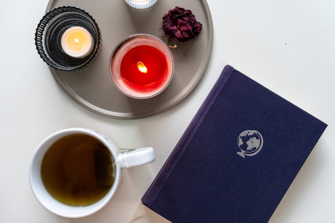 Man sieht ein blaues Notizbuch mit einem silbernen Globus drauf. Es liegt neben einer dunklen Tasse Tee und einem braunen Teller auf den eine rote Kerze, ein Teelicht und eine getrocknete Rose liegen.
