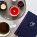 Man sieht ein blaues Notizbuch mit einem silbernen Globus drauf. Es liegt neben einer dunklen Tasse Tee und einem braunen Teller auf den eine rote Kerze, ein Teelicht und eine getrocknete Rose liegen.