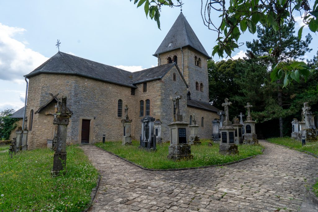 Im Mittelpunkt des Bildes steht eine alte Kirche. Im Vordergrund ist ein Weg zu sehen, der sich aufspaltet. Zwischen den wegen ist eine Rasenfläche, die mit alten Gräbsteinen gefüllt ist.