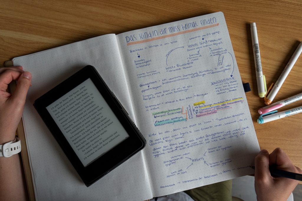 Auf einem Schreibtisch liegt ein Notizbuch mit einer vollgeschriebenen Seite. Diese trägt den Titel "Das Kind in dir muss Heimat finden". Auf der anderen hälfte des Notizbuches liegt ein geöffneter E-Reader. Man sieht zwei Hände. Eine Hält das Notizbuch, die andere schreibt. Daneben liegen vier bunte Textmarker.