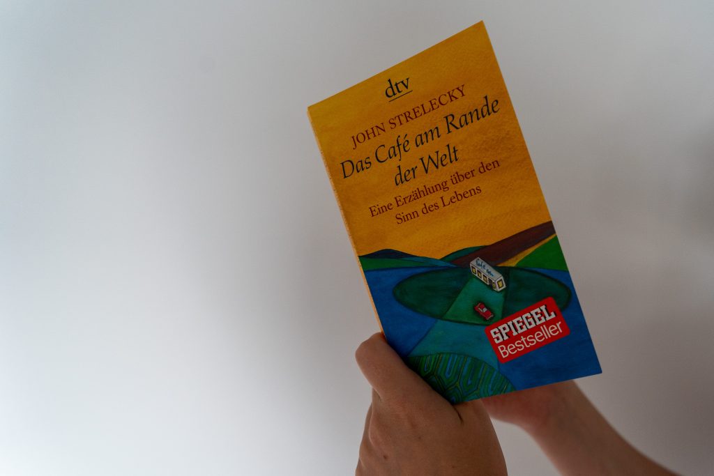Man sieht das Buch "Das Café am Rand der Welt" in einer Hand aufgeschlagen vor einem weißen Hintergrund