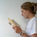 Auf dem Bild ist eine junge, blonde Frau. Sie trägt die Haare hochgesteckt und hat ein weißes T-Shirt in der Hand. Man sieht sie von der Seite. Sie hat ein offenes Buch in der Hand und scheint darin zu lesen.