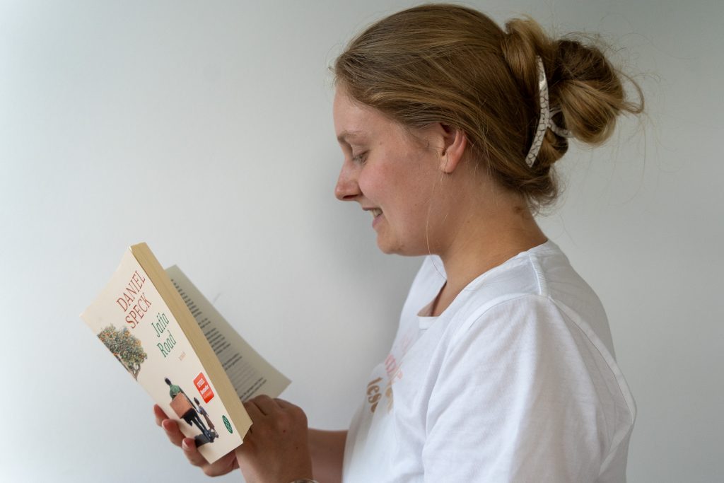 Auf dem Bild ist eine junge, blonde Frau. Sie trägt die Haare hochgesteckt und hat ein weißes T-Shirt in der Hand. Man sieht sie von der Seite. Sie hat ein offenes Buch in der Hand und scheint darin zu lesen. Sie lächelt dabei.