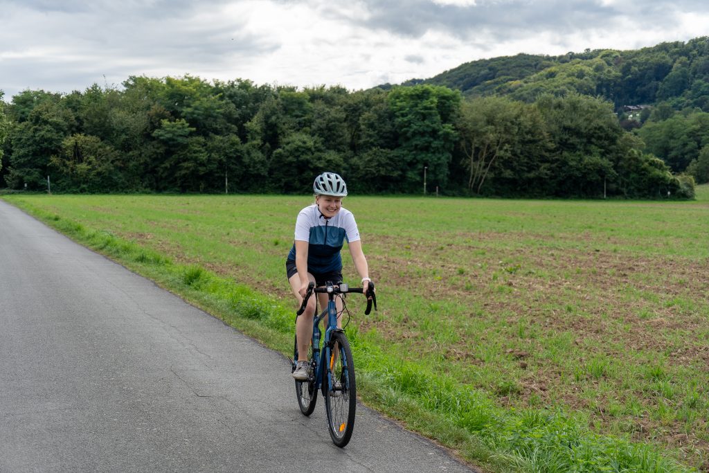 Auf dem Bild ist eine junge Frau in Radkleidung mit Helm zu sehen, die vor einer grünen Wiese auf einem Gravel-Bike fährt.