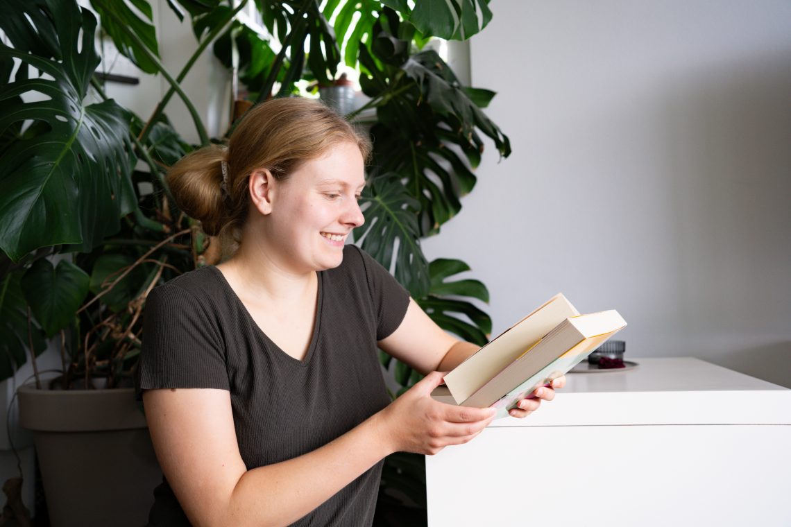 Man sieht eine Frau mit blonden Haaren in einem grünen Shirt. Sie sitzt vor einer Monstera und hält zwei Bücher in der Hand.