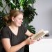 Man sieht eine Frau mit blonden Haaren in einem grünen Shirt. Sie sitzt vor einer Monstera und hält zwei Bücher in der Hand.